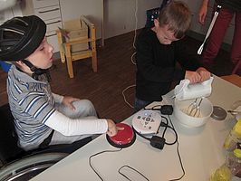 Zwei Jungen backen Kuchen. Der Junge im Rollstuhl betätigt dafür einen elektrischen Taster um das Handrührgerät in Gang zu bringen. Mit diesem rührt der andere Junge den Teig.
